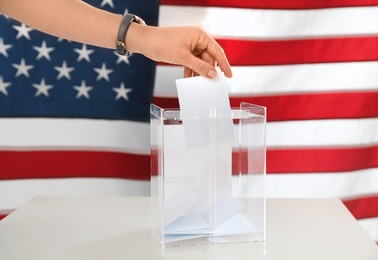 Woman putting ballot paper into box and American flag on background, closeup