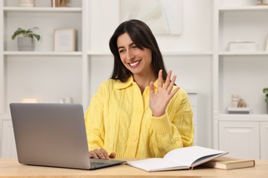 Photo of Happy woman waving hello during video chat via laptop at table indoors