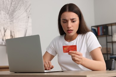 Photo of Confused woman with credit card using laptop at table indoors. Be careful - fraud