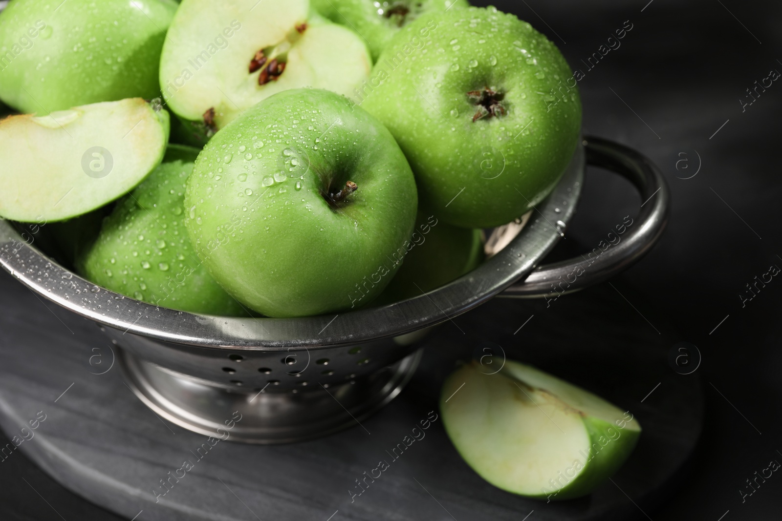 Photo of Ripe green apples with water drops and colander on black table, closeup