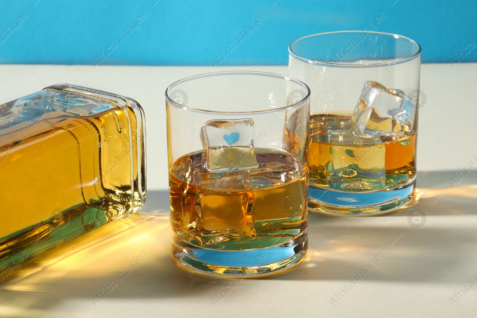 Photo of Whiskey with ice cubes in glasses and bottle on white table, closeup