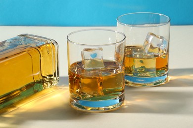 Whiskey with ice cubes in glasses and bottle on white table, closeup