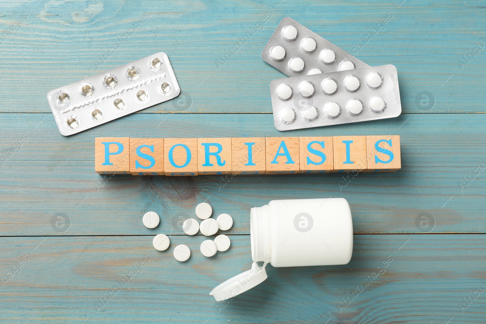 Photo of Word Psoriasis made of cubes with letters and pills on light blue wooden table, flat lay