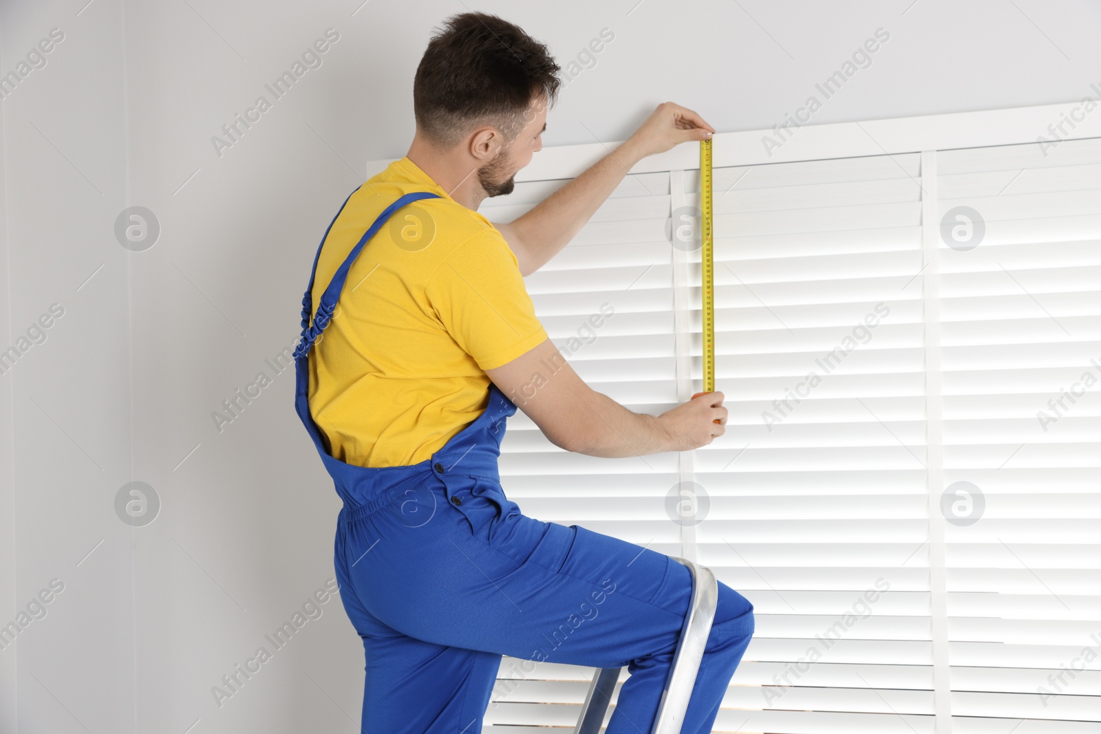 Photo of Worker in uniform using measuring tape while installing horizontal window blinds indoors