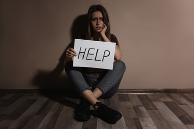 Abused young woman with sign HELP near beige wall. Domestic violence concept