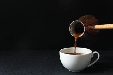 Photo of Turkish coffee. Pouring brewed beverage from cezve into cup on table against black background, space for text