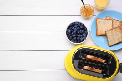 Yellow toaster with roasted bread, glass of juice, blueberries and jam on white wooden table, flat lay. Space for text