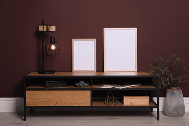Empty frames and stylish lamp on wooden table near brown wall. Mockup for design