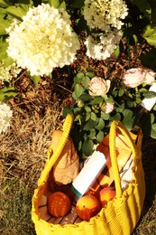 Yellow wicker bag with roses, peaches, baguette and wine outdoors, above view