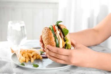 Woman holding tasty burger over plate at table, closeup. Space for text