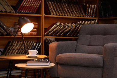Photo of Book with glasses, table lamp and cup of drink near comfortable armchair in cozy home library
