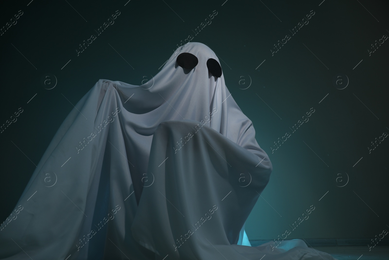 Photo of Creepy ghost. Woman covered with sheet on dark teal background, low angle view. Space for text