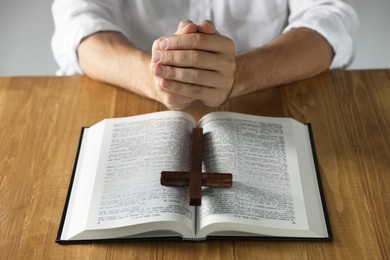 Man with Bible and cross praying at wooden table, closeup