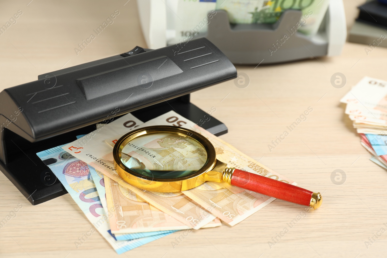 Photo of Modern currency detector with Euro banknotes and magnifying glass on wooden table. Money examination device