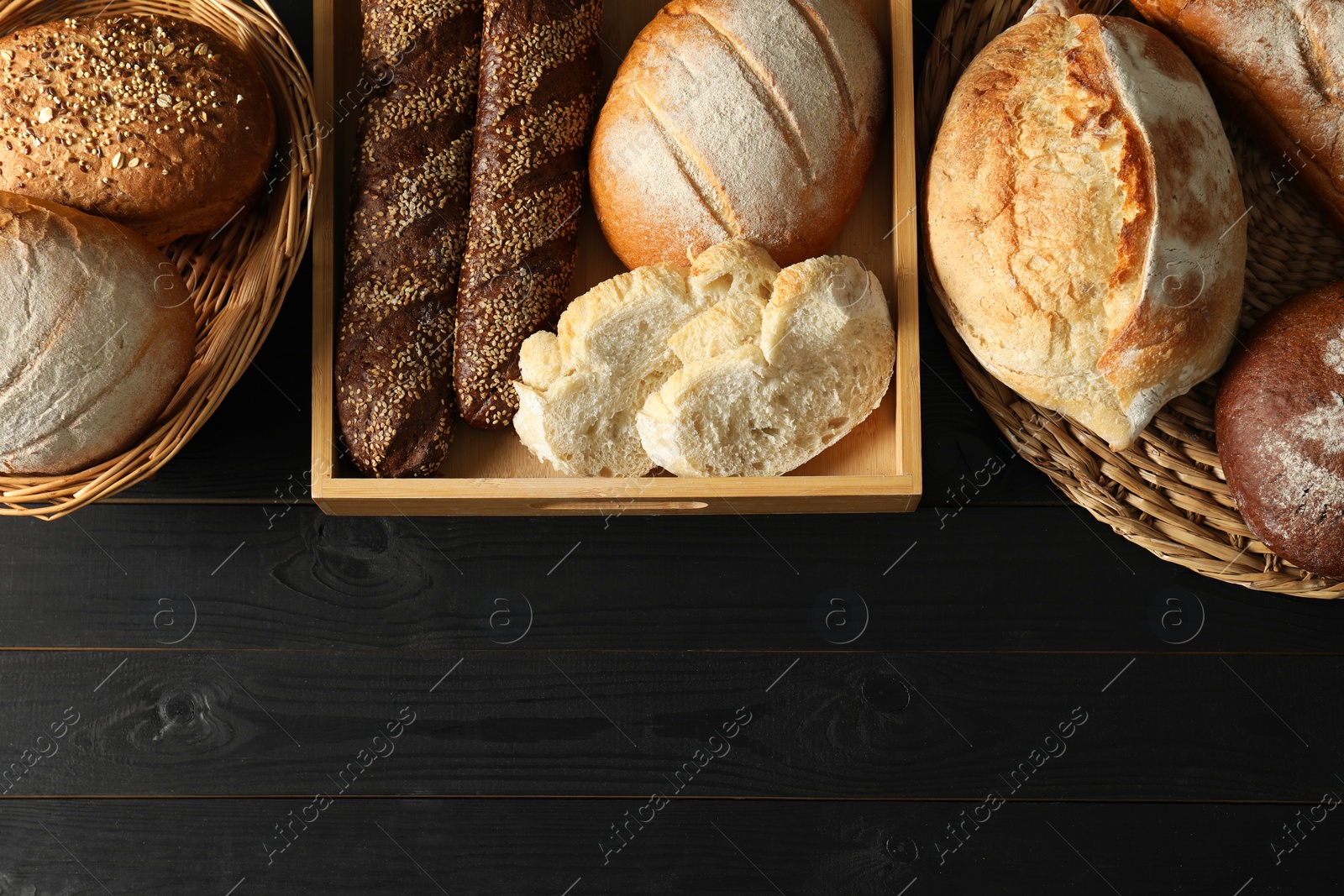 Photo of Wicker baskets with different types of fresh bread on black wooden table, flat lay. Space for text