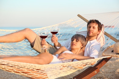 Photo of Young couple resting with glasses of wine in hammock on beach