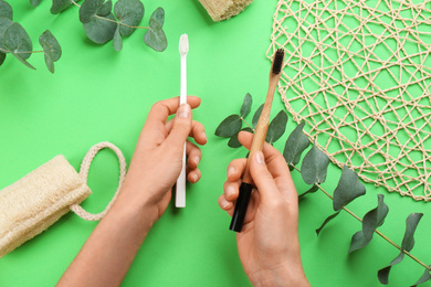 Photo of Woman holding natural bamboo and plastic toothbrushes over green background, top view