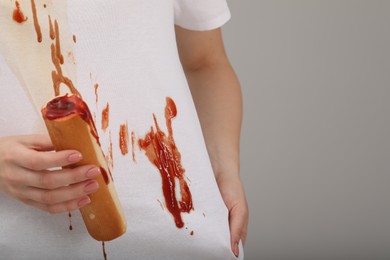 Woman holding hotdog and showing stain from sauce on her shirt against light grey background, closeup. Space for text