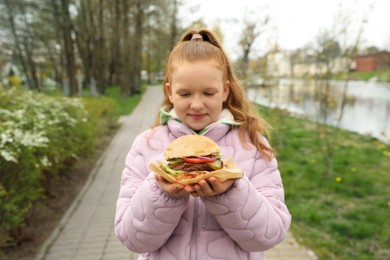 Photo of Little girl holding fresh delicious burger outdoors. Street food