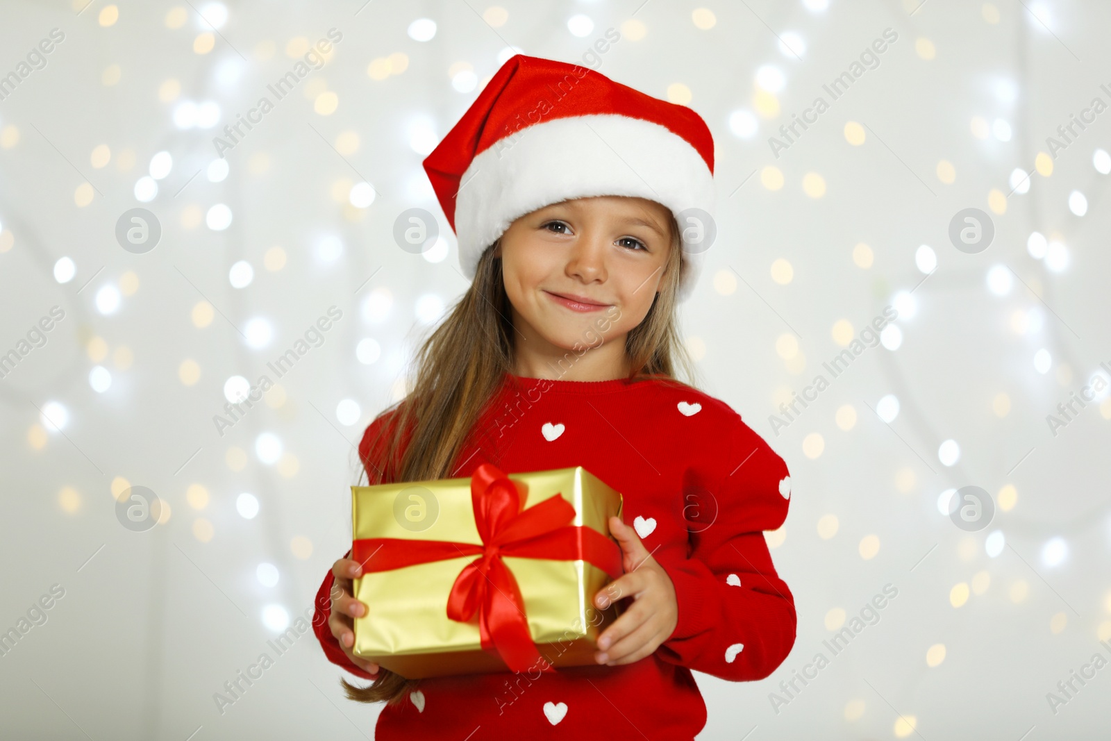 Photo of Happy little child in Santa hat with gift box against blurred festive lights. Christmas celebration