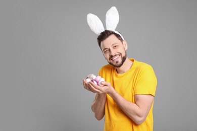 Photo of Happy man in bunny ears headband holding painted Easter eggs on grey background. Space for text
