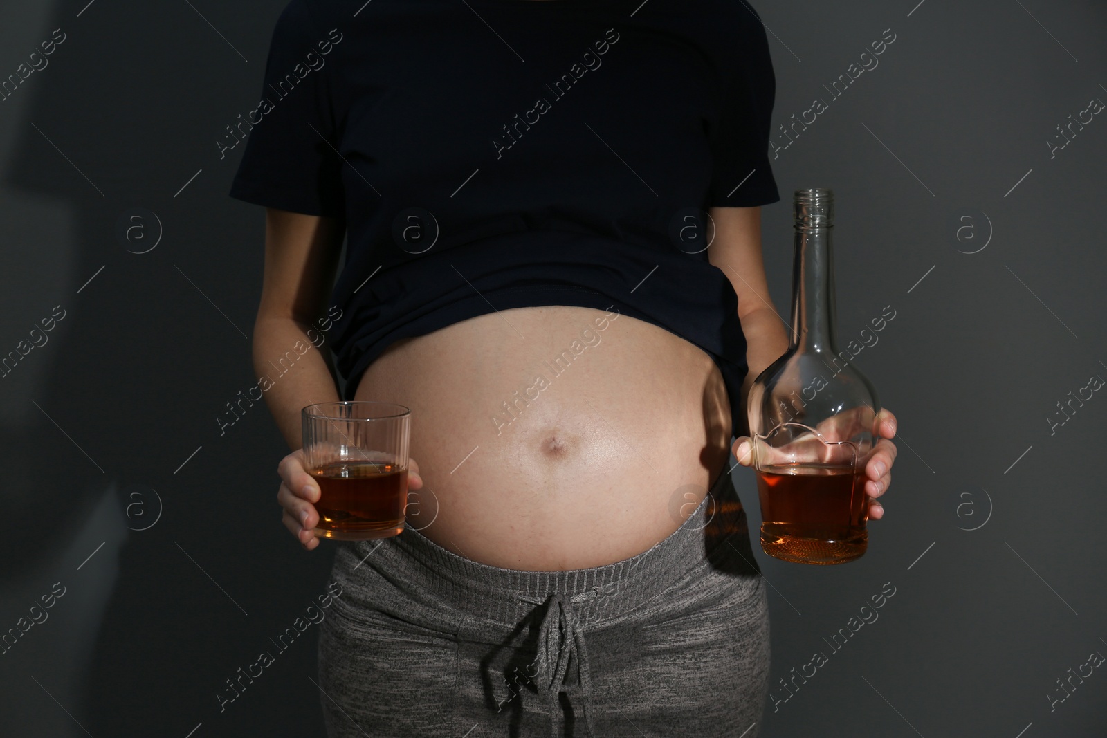 Photo of Pregnant woman with glass and bottle of whiskey on dark background. Alcohol addiction