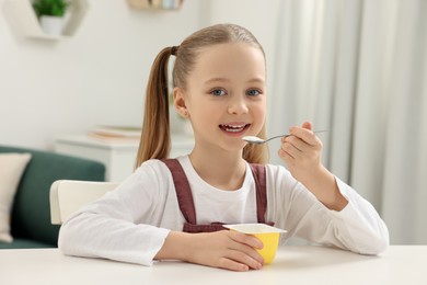 Cute little girl with tasty yogurt at white table indoors