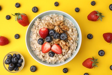 Photo of Tasty oatmeal with strawberries, blueberries and walnuts in bowl surrounded by ingredients on yellow background, flat lay