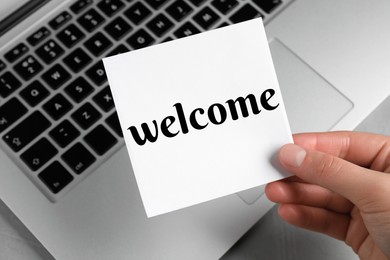 Woman holding paper note with word Welcome over laptop, closeup