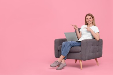 Photo of Happy woman with laptop sitting in armchair and pointing at something on pink background. Space for text