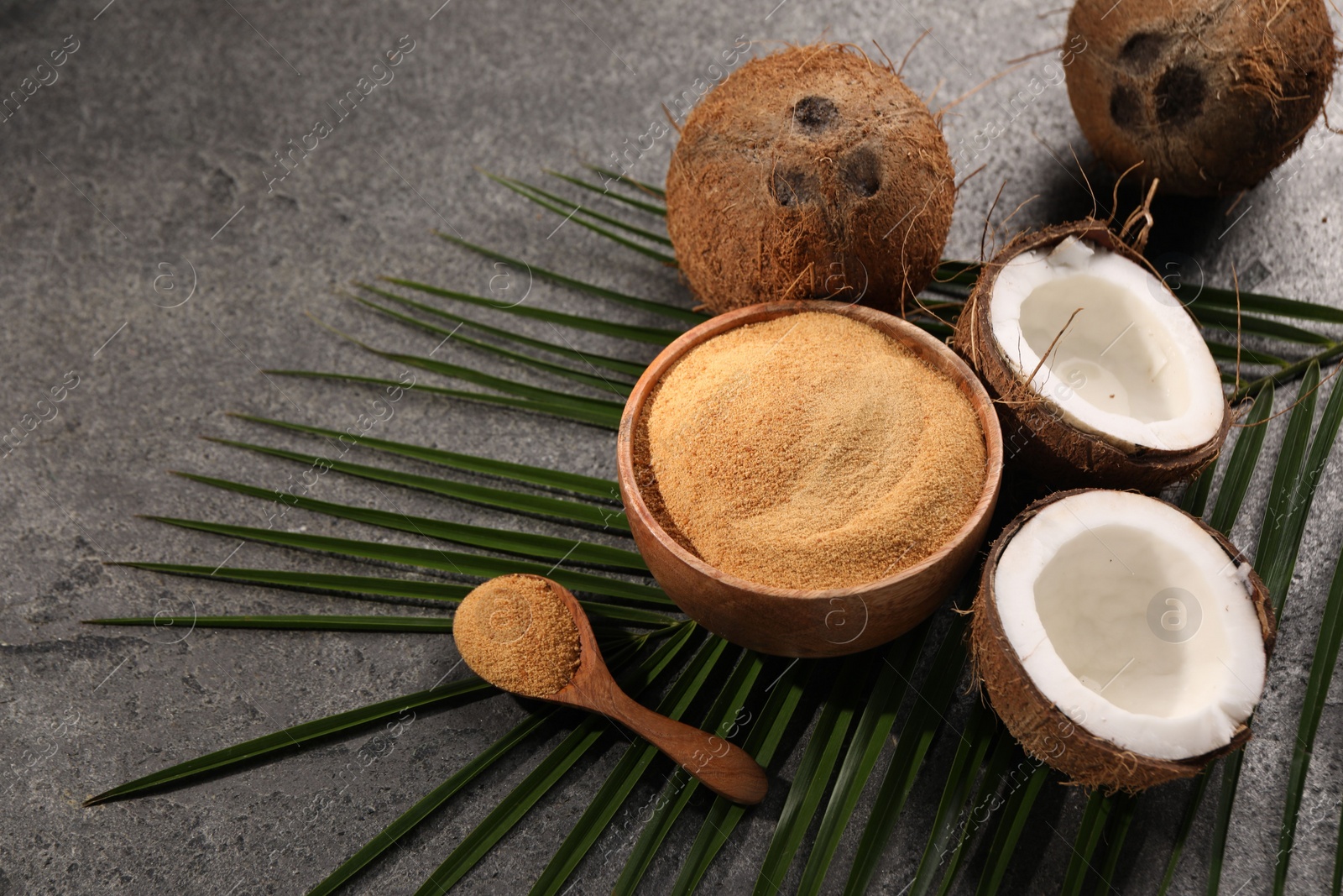 Photo of Coconut sugar in bowl, spoon, palm leaves and fruits on dark textured table, closeup. Space for text