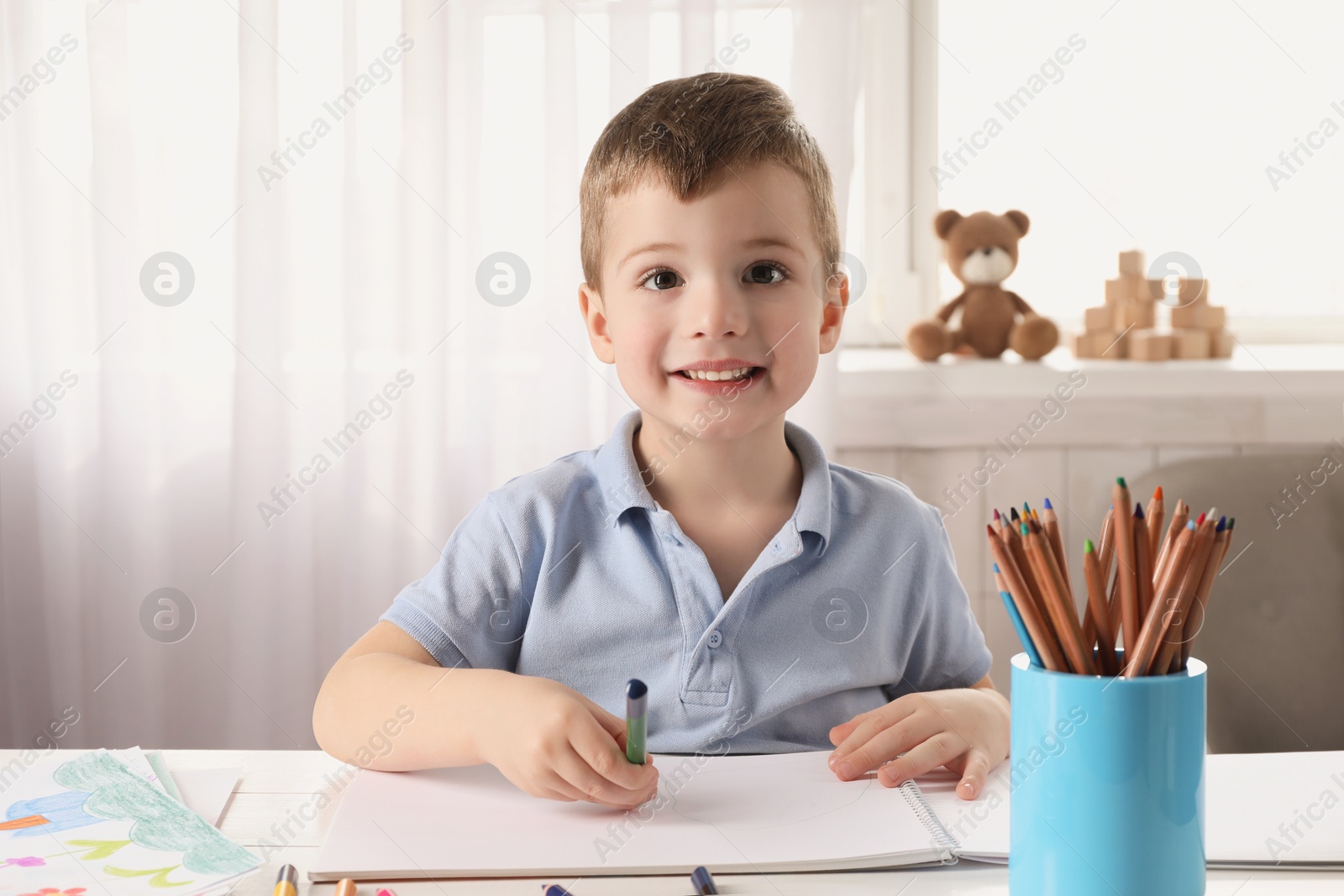 Photo of Cute little boy drawing with pencil at white table in room. Child`s art