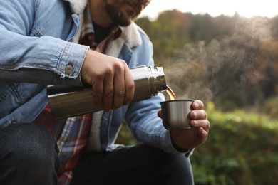 Man pouring hot drink from metallic thermos into cup lid in nature, closeup