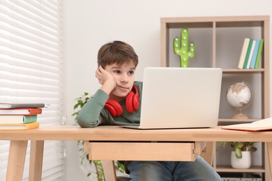 Photo of Tired boy with red headphones using laptop at desk in room. Home workplace