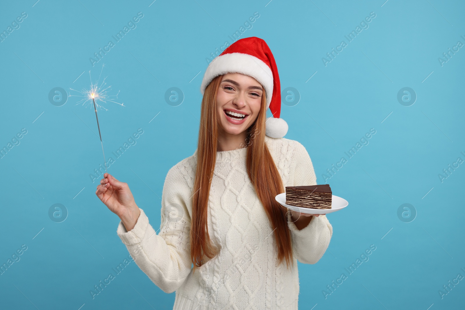 Photo of Young woman in Santa hat with piece of tasty cake and burning sparkler on light blue background