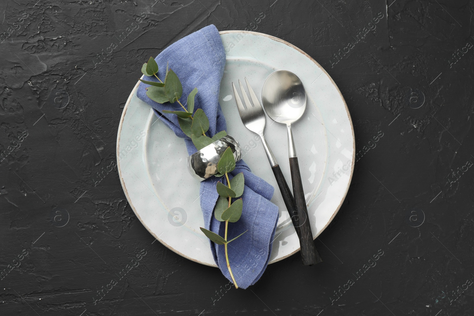 Photo of Stylish setting with cutlery, eucalyptus branch, napkin and plate on dark textured table, top view