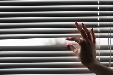 Photo of Woman separating slats of white blinds indoors, closeup