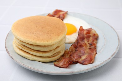 Photo of Plate with tasty pancakes, fried egg and bacon on white tiled table, closeup