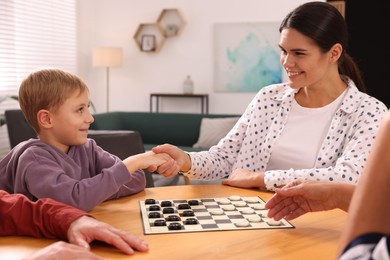 Photo of Family playing checkers at wooden table in room. Mother and son shaking hands after game