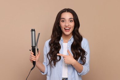 Photo of Excited woman pointing at curling hair iron on beige background