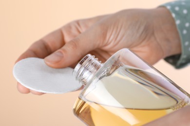 Photo of Woman pouring micellar water from bottle on cotton pad against beige background, closeup