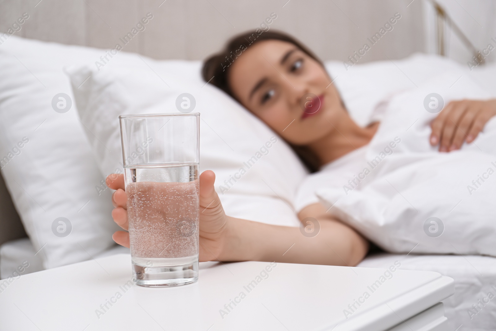 Photo of Young woman taking glass of water from nightstand at home, focus on hand