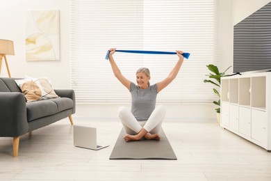 Photo of Senior woman doing exercise with fitness elastic band near laptop on mat at home