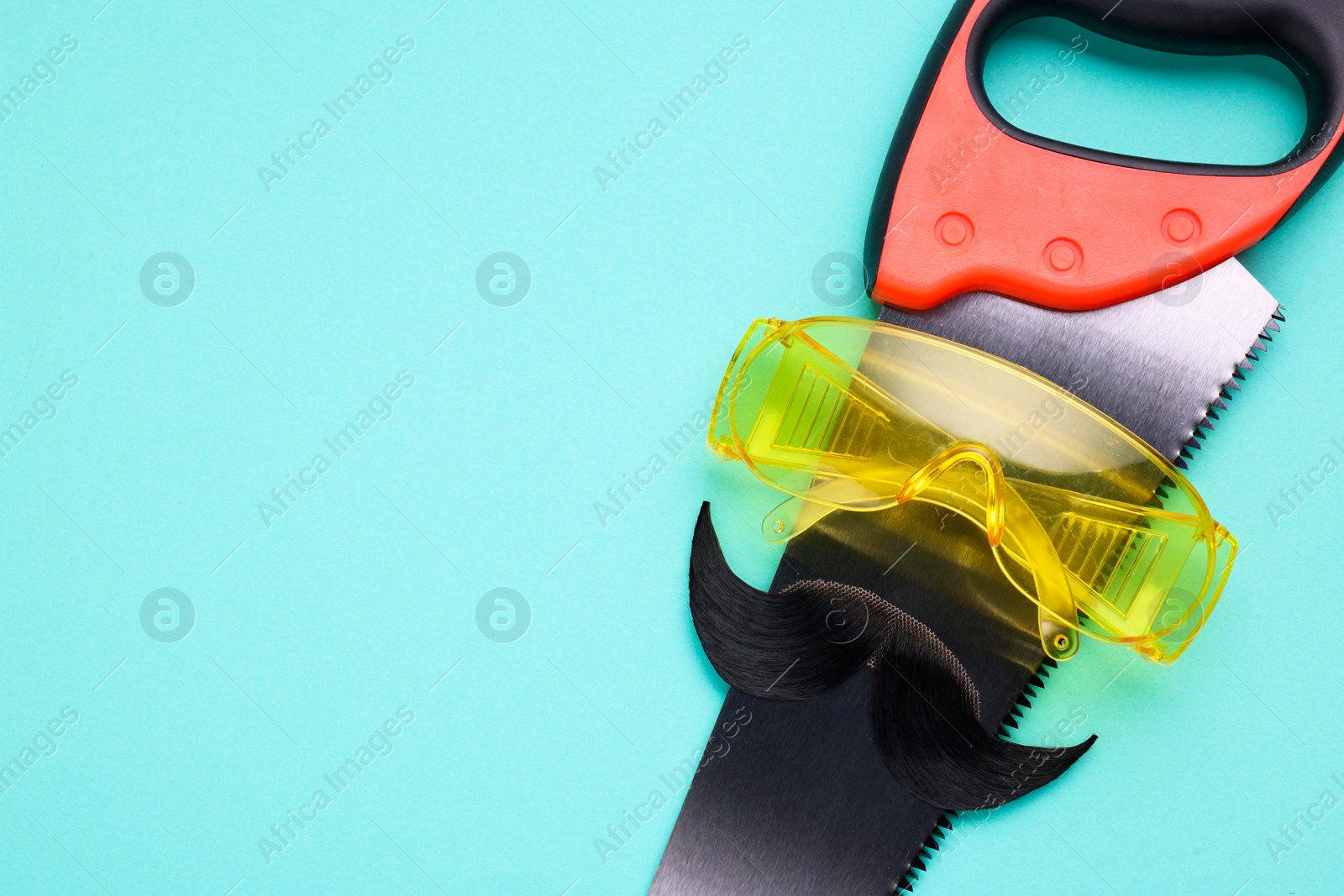 Photo of Man's face made of artificial mustache, safety glasses and hand saw on light blue background, top view. Space for text