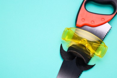 Man's face made of artificial mustache, safety glasses and hand saw on light blue background, top view. Space for text