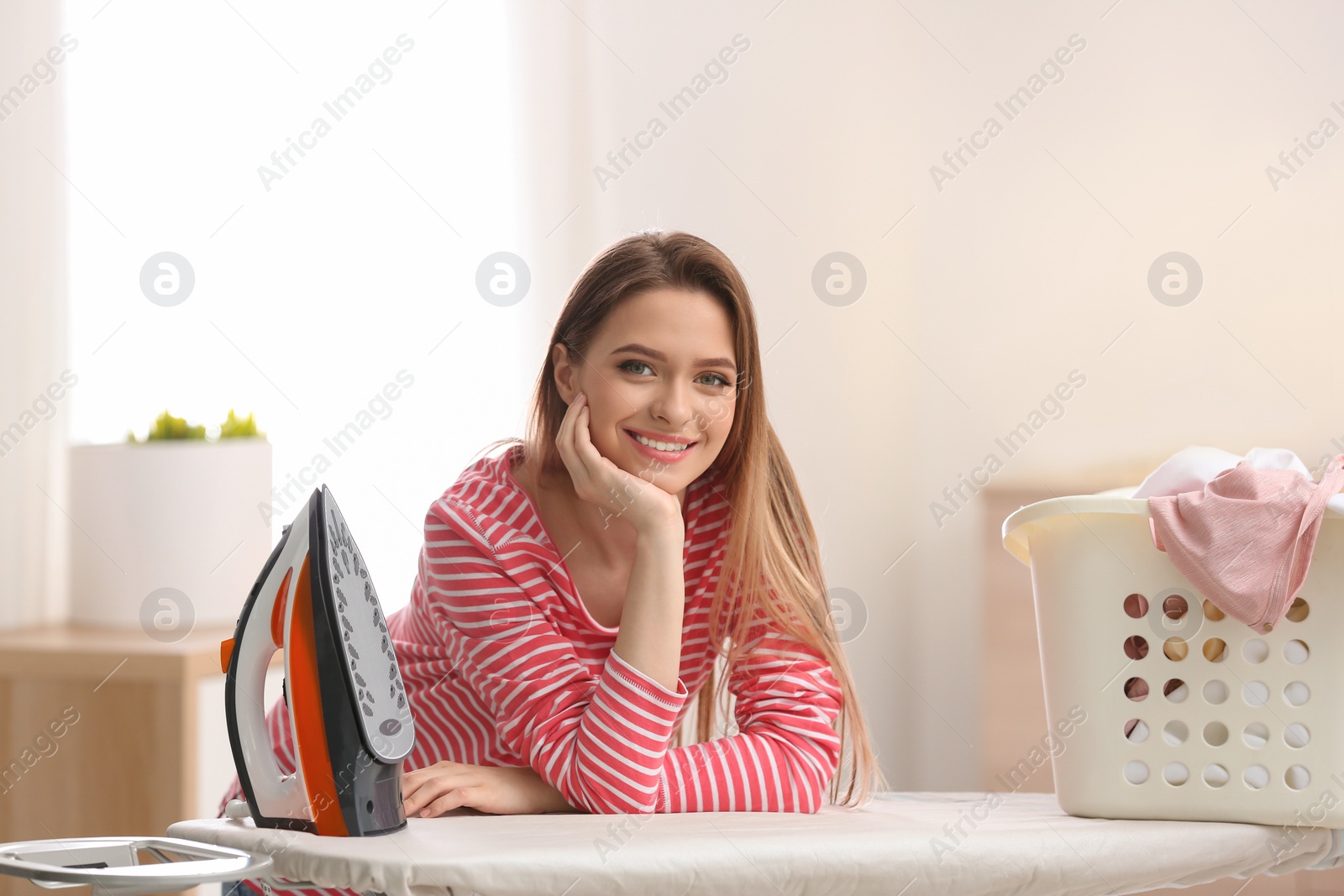 Photo of Young pretty woman with iron and basket of clean laundry at board indoors