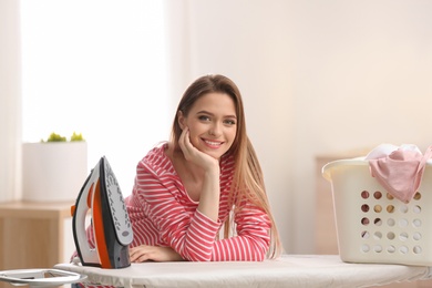 Young pretty woman with iron and basket of clean laundry at board indoors