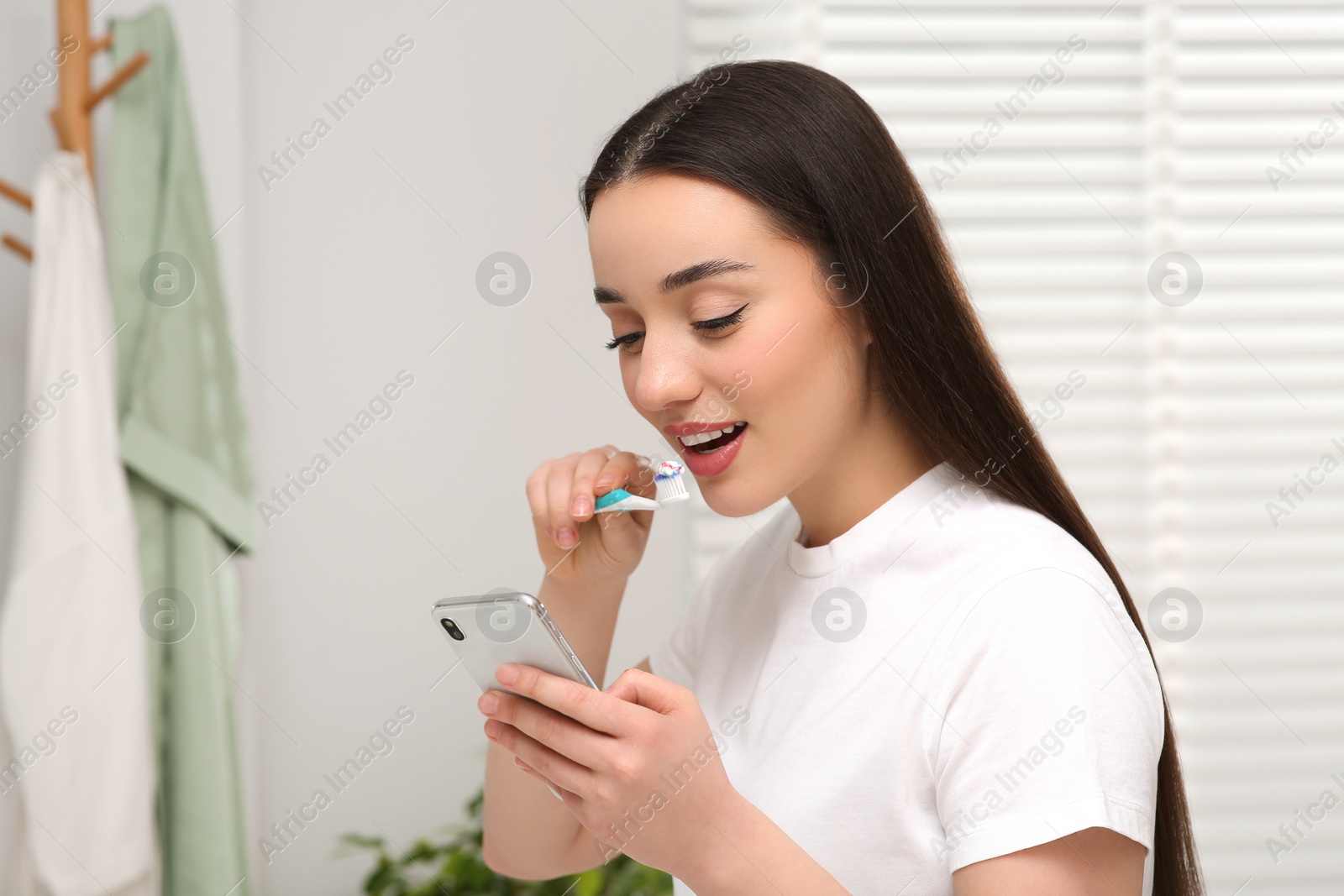 Photo of Beautiful young woman using smartphone while brushing teeth in bathroom. Internet addiction