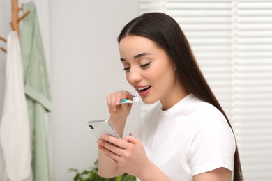 Photo of Beautiful young woman using smartphone while brushing teeth in bathroom. Internet addiction