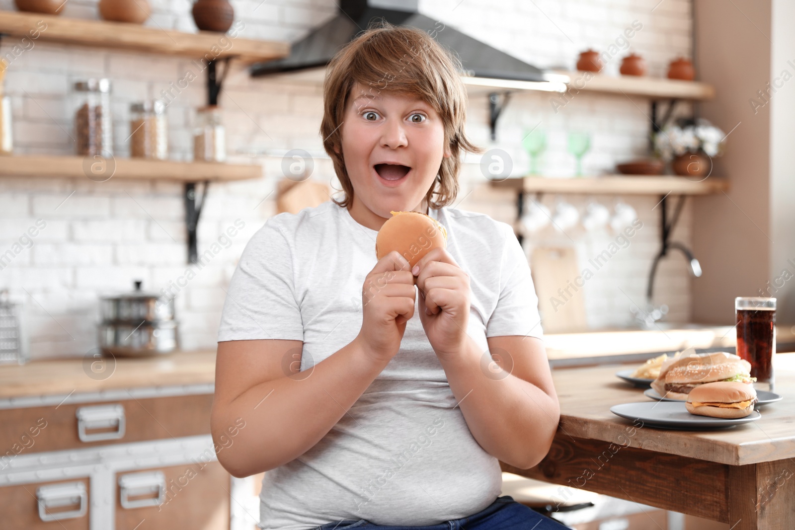 Photo of Emotional overweight boy at table with fast food in kitchen